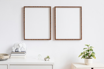Elegant home interior with two brown wooden mock up photo frames above the white shelf with books, air plants, wooden basket and geometric elephant figure. Modern concept of white room decor. 