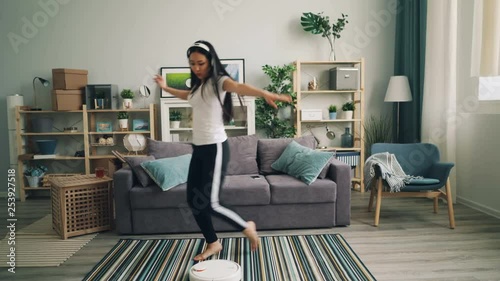 Asian young lady is enjoyig modern technology listening to music in wireless headphones and dancing while robotic hoover is cleaning floor in her nice apartment. photo