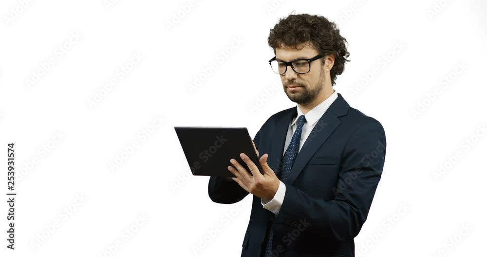 Caucasian man in glasses, suit and tie standing on the white background and typing on the laptop, then smiling and posing to the camera. Caucasian man in glasses, suit and tie standing on the white background and typing on the laptop, then smiling and posing to the camera.