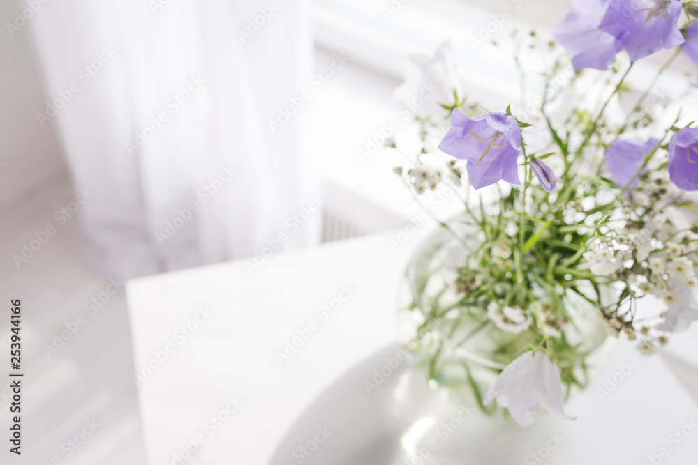 Glass vase with lilac and white floweers  in light cozy bedroom interior. White wall, sunlight from window, copy space