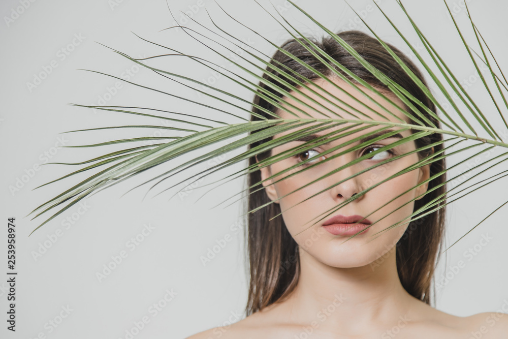 Fototapeta premium An attractive happy girl smiles wide and covers half of her face with fern leaves from the sun. Green, healthy skin and eco-friendly concept.