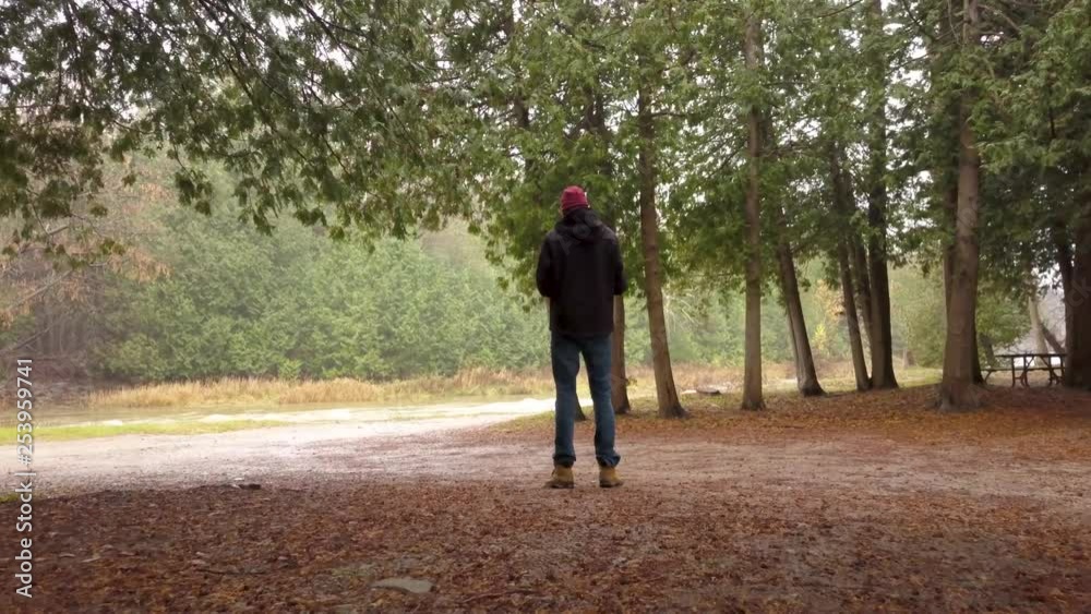 Coming up behind a man in a black coat under some trees in a park near a river. Coming up behind a man in a black coat under some trees in a park near a river.