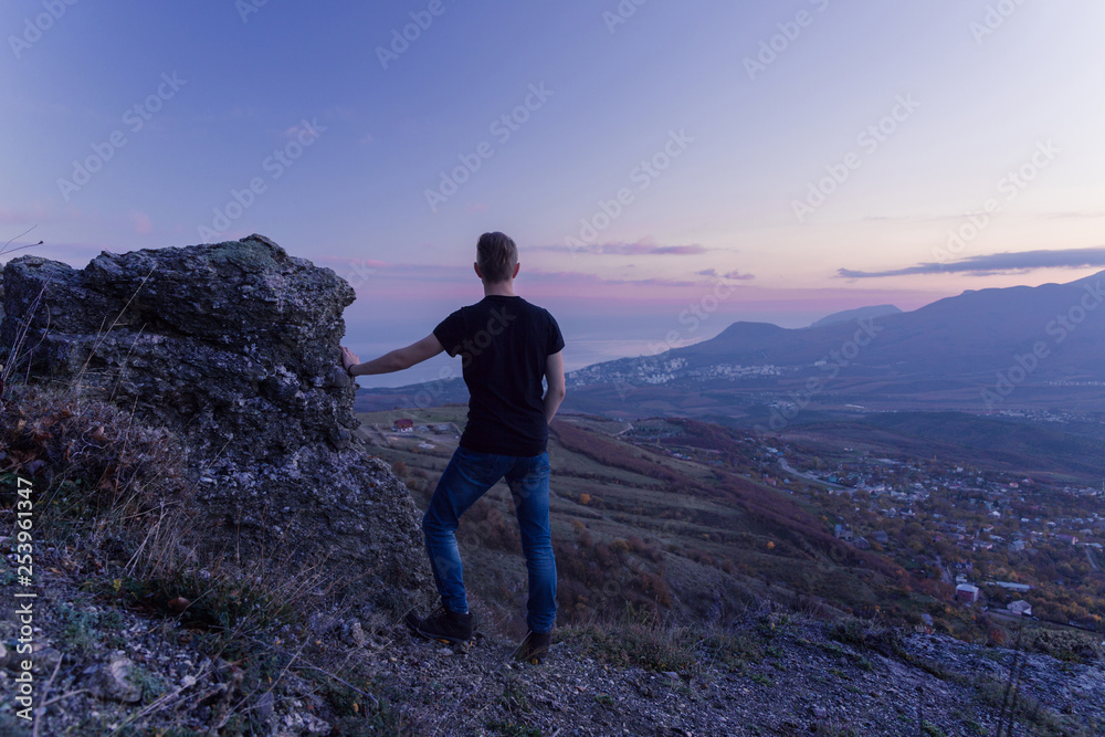 A young man in a black T-shirt stands high in the mountains and looks at a purple sunset.