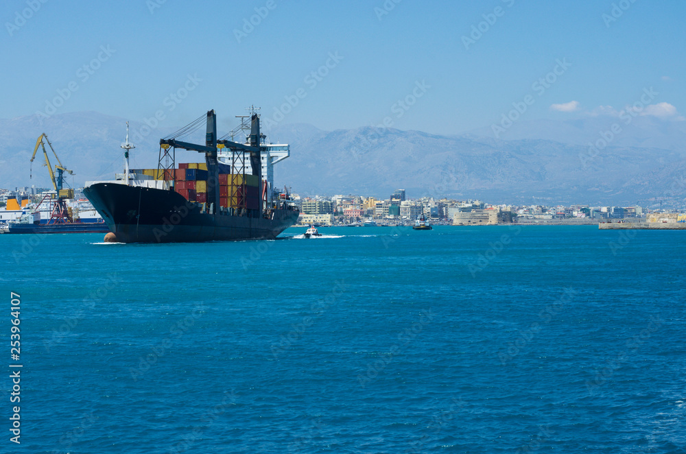 Container ship sailing from the Bay of Heraklion, accompanied by a pilot ship in the background of the mountains and the port