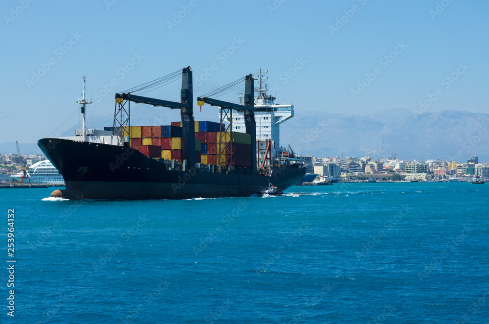 Container ship sailing from the Bay of Heraklion, accompanied by a pilot ship in the background of the mountains and the port