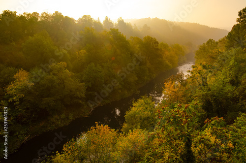 Magical sunrise over deep foggy autumn forest along a river. First rays of sun through fog and trees on slopes. Scenic park fall landscape of misty old forest with sun rays, shadows and fog.