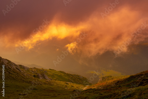 Natural alpine scenery landscape of mountains and dramatic foggy clouds on fire, strong orange color from the rising sun. High altitude orange clouds background at sunrise.