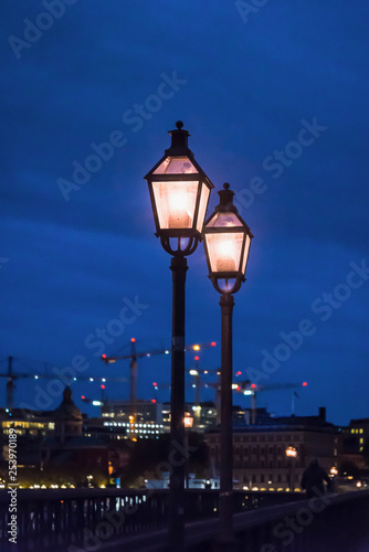 Illuminated street lanterns at night and construction site cranes in the background, Stockholm, Sweden © Marina Marr