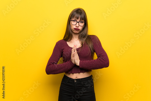 Woman with glasses over yellow wall keeps palm together. Person asks for something © luismolinero