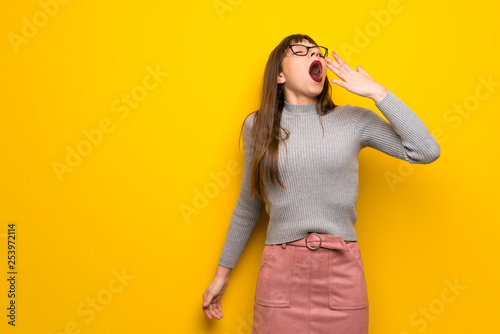Woman with glasses over yellow wall yawning and covering wide open mouth with hand