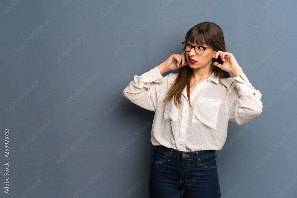 Woman with glasses over blue wall covering both ears with hands Woman with glasses over blue wall covering both ears with hands