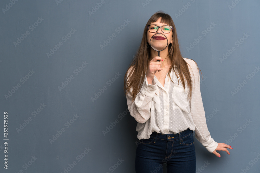 Woman with glasses over blue wall taking a magnifying glass and showing teeth through it Woman with glasses over blue wall taking a magnifying glass and showing teeth through it