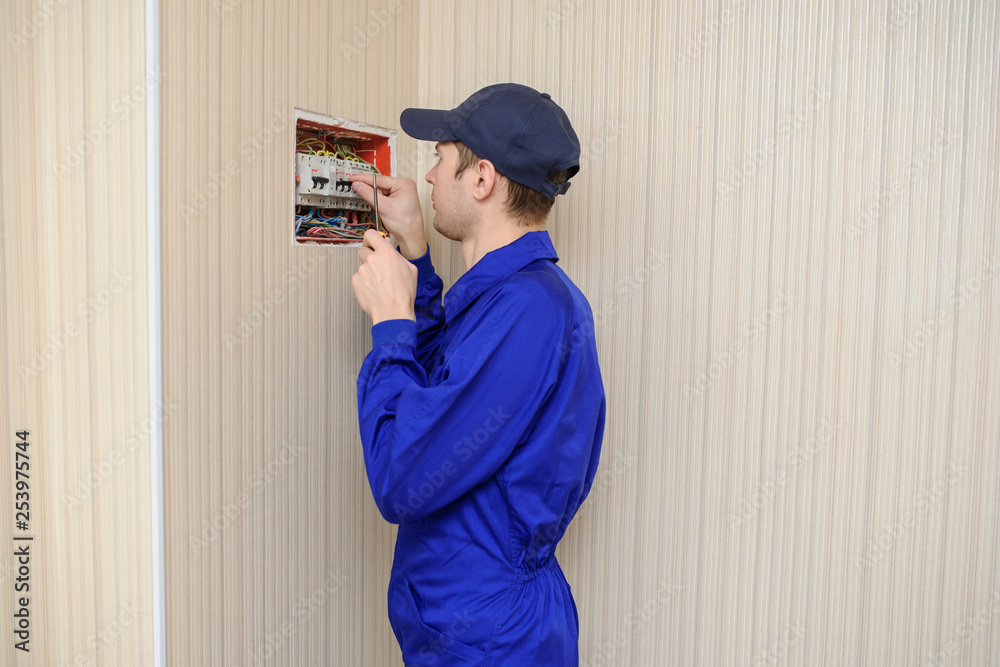 lateral view of a young eletrician in blue overall disassembling a electrical panel with fuses in a house. lateral view of a young eletrician in blue overall disassembling a electrical panel with fuses in a house.