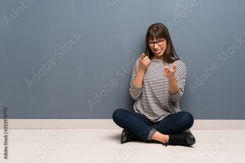 Woman with glasses sitting on the floor pointing to the front and smiling