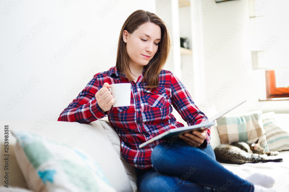 Naklejka premium Young woman reading book on sofa relaxing at home