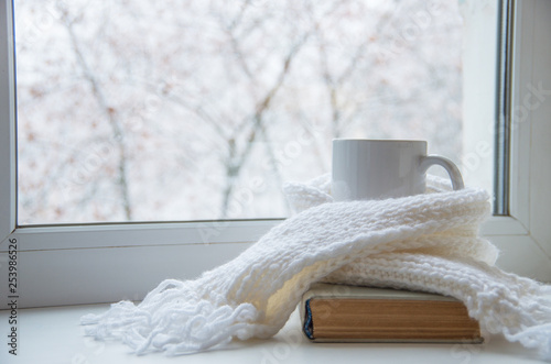 Fototapeta Mug of hot tea and warm woolen knitting on windowsill against snow landscape from outside