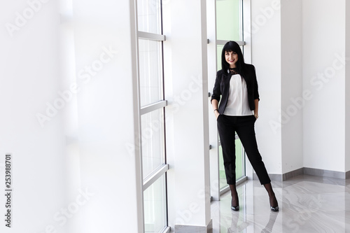 Young Attractive happy brunette woman dressed in a black business suit standing near window in a office, smiling, looking at camera.