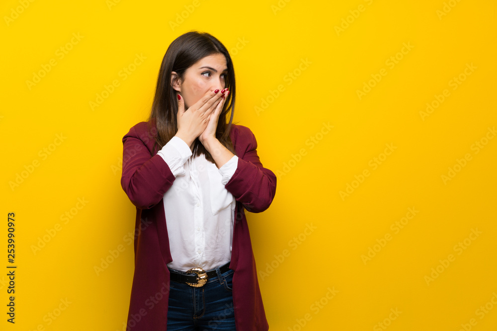 Young woman over yellow wall covering mouth and looking to the side Young woman over yellow wall covering mouth and looking to the side