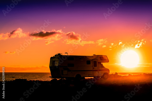 Amazing view of a camper parking near the sea on a summer vacation day at the sunset. Beautiful holiday landscape of a caravan with ocean and sun in background. Orange light of a sunrise with clouds.