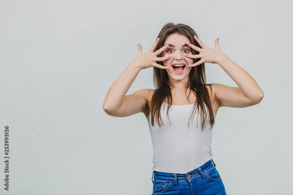 A pretty young girl standing on a white background. The girl is dressed in a white shirt. Has beautiful black long hair. Having put all the fingers of two hands, the elongated girl puts them on the