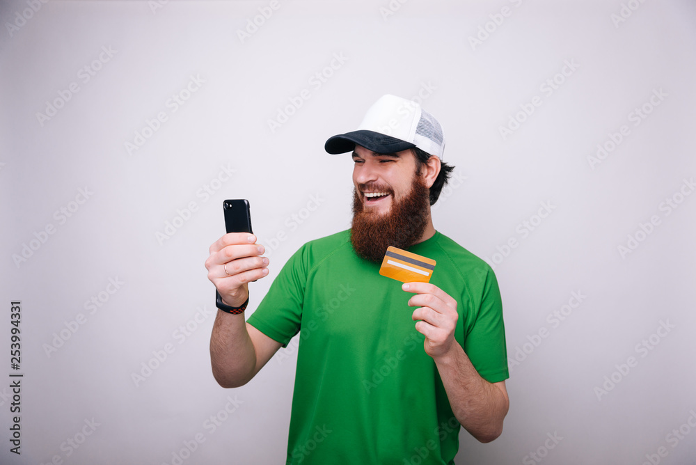 Image of happy cheerful bearded man standing isolated over liight gray wall background holding credit card using mobile phone.