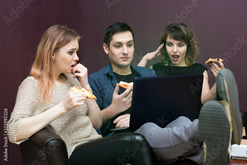 Young people eating pizza at the laptop. A man and two women are watching a movie on a computer.