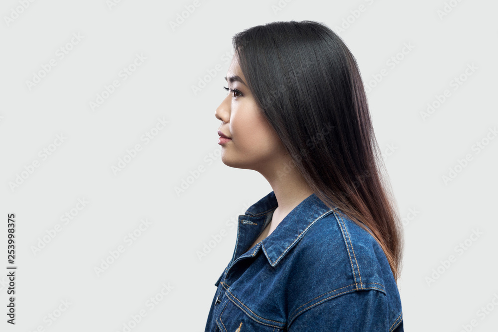 Profile side view portrait of calm serious beautiful brunette asian young woman in casual blue denim jacket with makeup standing and looking. indoor studio shot, isolated on light grey background. Profile side view portrait of calm serious beautiful brunette asian young woman in casual blue denim jacket with makeup standing and looking. indoor studio shot, isolated on light grey background.