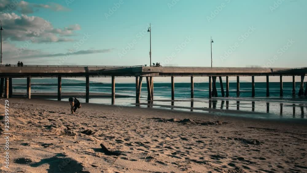 Slow zoom in fast timelapse low angle view of section of Santa Monica pier, Venice beach. Fast motion people passing along sandy beach. Slow zoom in fast timelapse low angle view of section of Santa Monica pier, Venice beach. Fast motion people passing along sandy beach.