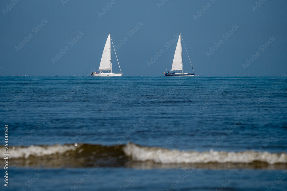 Two sailboats with white sails coasting in deep blue water not far from the beach on a sunny day