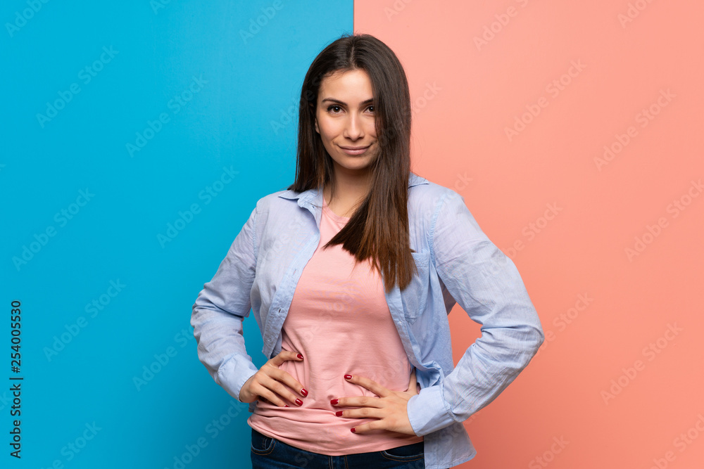 Young woman over pink and blue wall posing with arms at hip and laughing looking to the front Young woman over pink and blue wall posing with arms at hip and laughing looking to the front