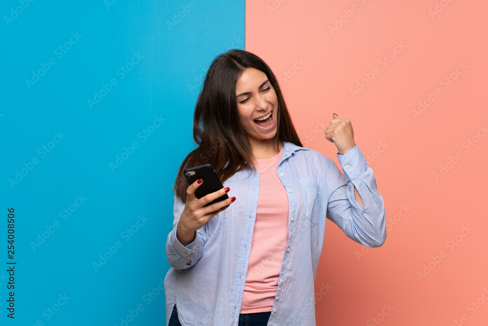Young woman over pink and blue wall celebrating a victory with a mobile