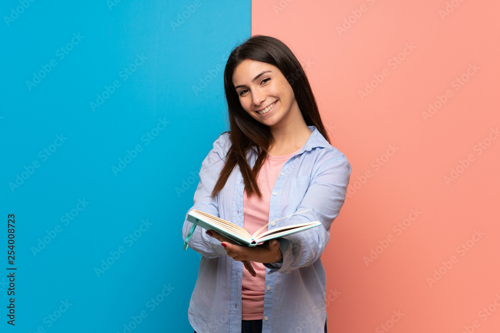 Young woman over pink and blue wall holding a book and giving it to someone Young woman over pink and blue wall holding a book and giving it to someone