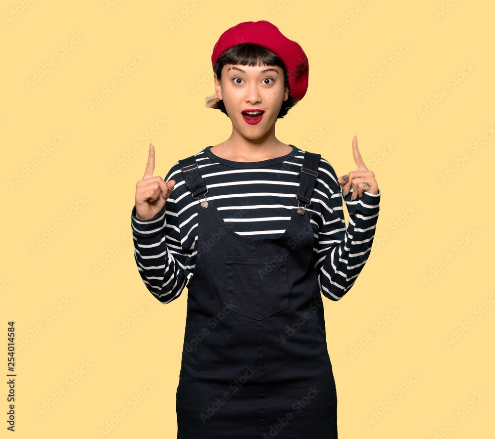 Young woman with beret pointing up and surprised over yellow background Young woman with beret pointing up and surprised over yellow background