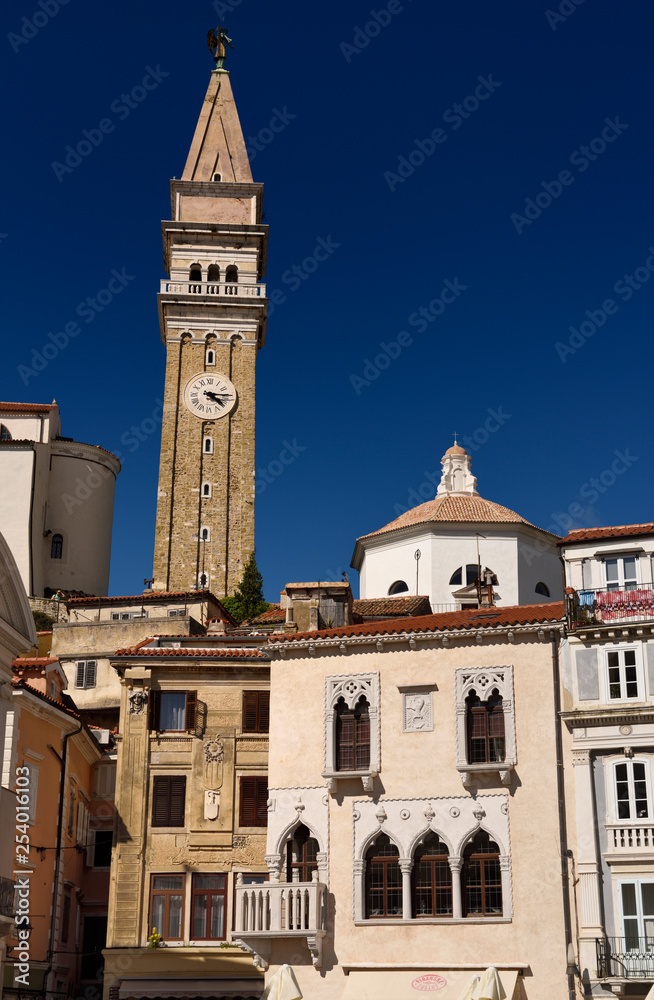 Fototapeta premium Bell and clock tower of St George's Parish Catholic Cathedral and baptistery with pink Venetian House in Tartini Square Piran Slovenia