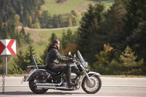 Side view of bearded long-haired motorcyclist in sunglasses and black leather clothing riding cruiser motorbike along narrow asphalt path on sunny day on background of tall trees and greeen grass.