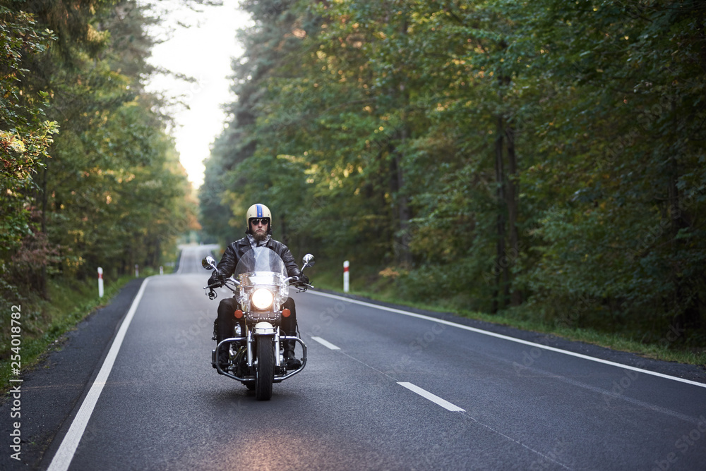 Bearded biker in sunglasses, helmet and black leather clothing riding cruiser motorcycle along asphalt road winding among tall green trees on sunny summer evening. Bearded biker in sunglasses, helmet and black leather clothing riding cruiser motorcycle along asphalt road winding among tall green trees on sunny summer evening.