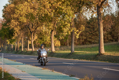 Bearded biker in sunglasses and black leather clothing riding modern powerful motorcycle along empty asphalt country road by beautiful tall trees with golden foliage on sunny autumn day.