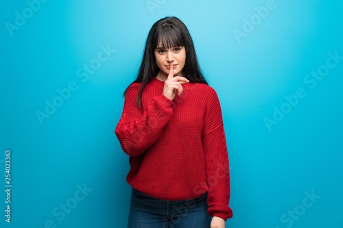 Woman with red sweater over blue wall showing a sign of silence gesture putting finger in mouth