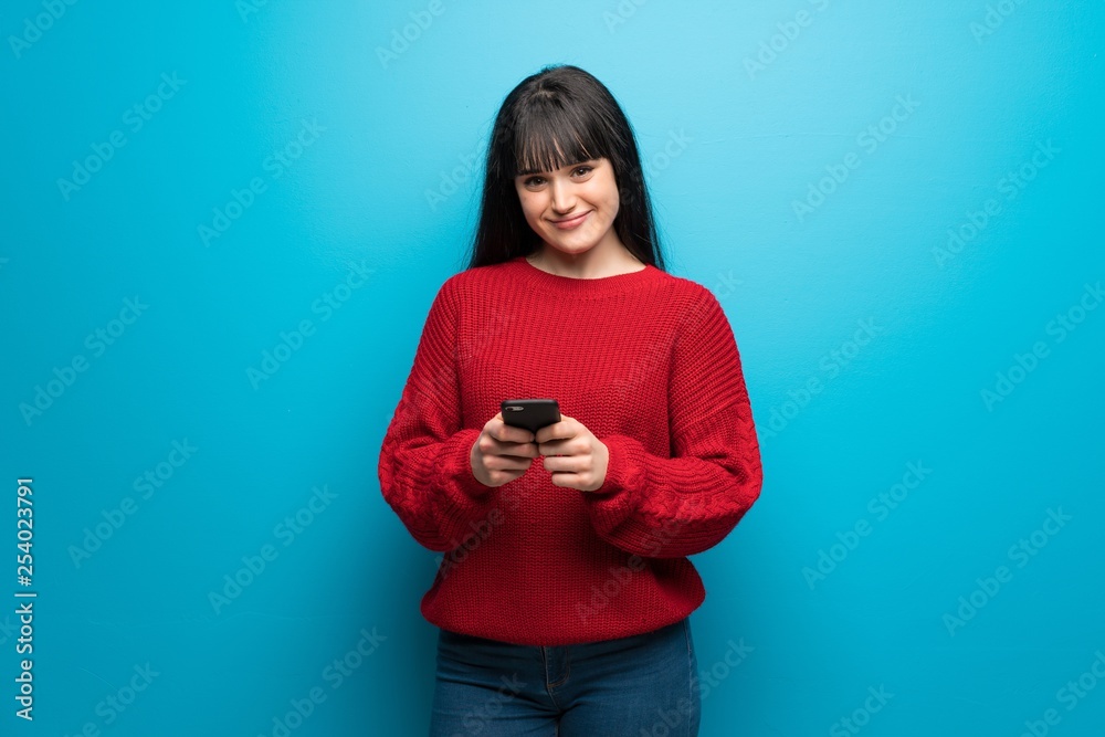 Woman with red sweater over blue wall sending a message with the mobile Woman with red sweater over blue wall sending a message with the mobile