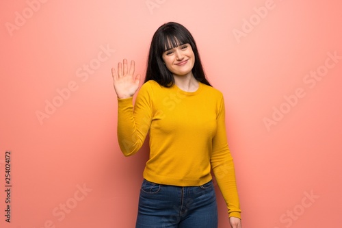 Woman with yellow sweater over pink wall saluting with hand with happy expression