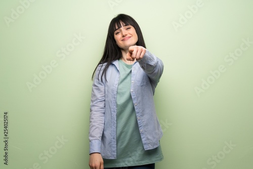 Young woman over green wall points finger at you with a confident expression