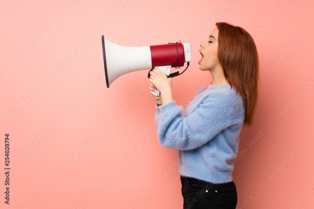 Young redhead woman over pink background shouting through a megaphone Young redhead woman over pink background shouting through a megaphone