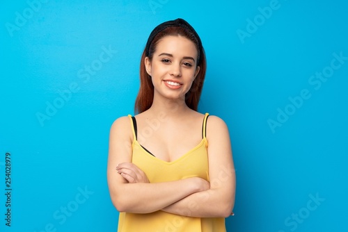 Young redhead woman over blue background keeping the arms crossed in frontal position