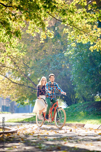 Young active smiling traveler couple, handsome bearded man and attractive blond woman riding together tandem double bicycle along path in lit by morning sun beautiful autumn park under tall trees.