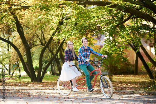 Young tourist couple, handsome bearded man and attractive blond long-haired woman cycling together tandem double bike by sunny alley with golden leaves on tall trees and hidden buildings background.