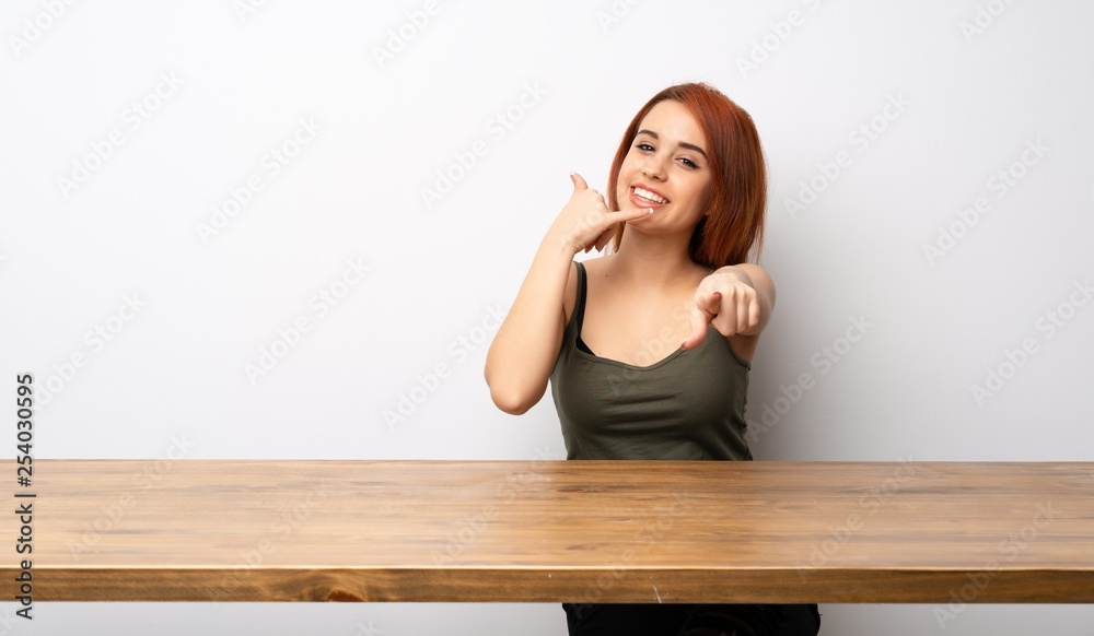 Young redhead woman at desk making phone gesture and pointing front Young redhead woman at desk making phone gesture and pointing front