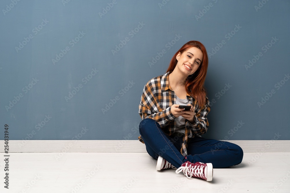 Naklejka premium Young redhead woman sitting on floor sending a message with the mobile