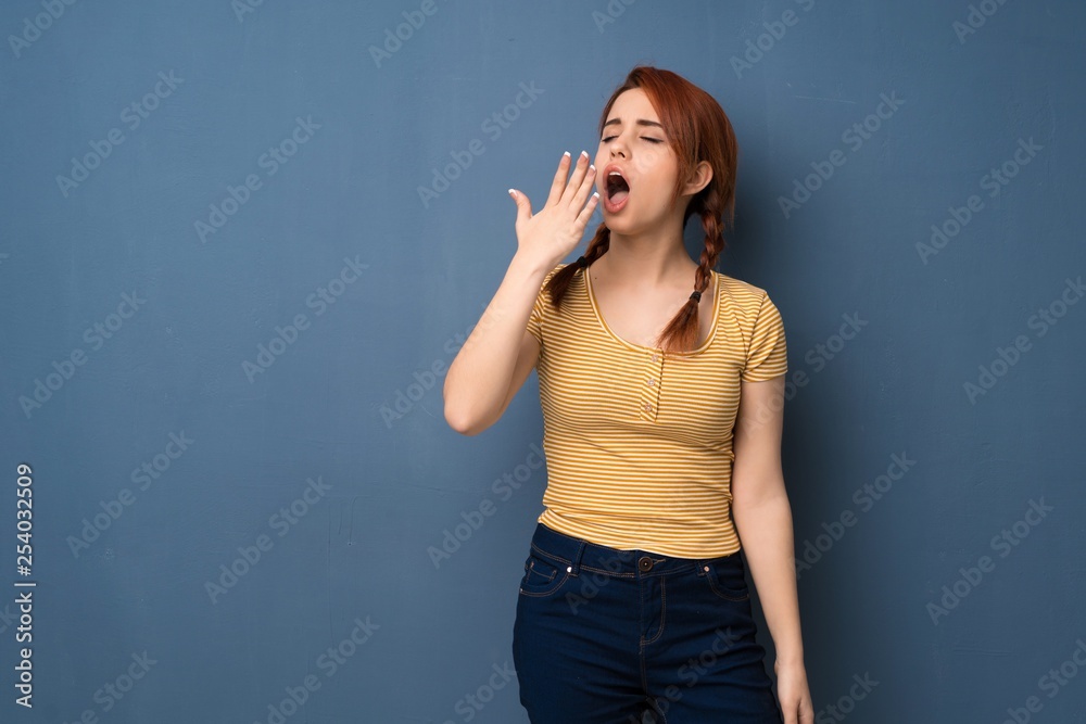 Young redhead woman over blue background yawning and covering wide open mouth with hand Young redhead woman over blue background yawning and covering wide open mouth with hand