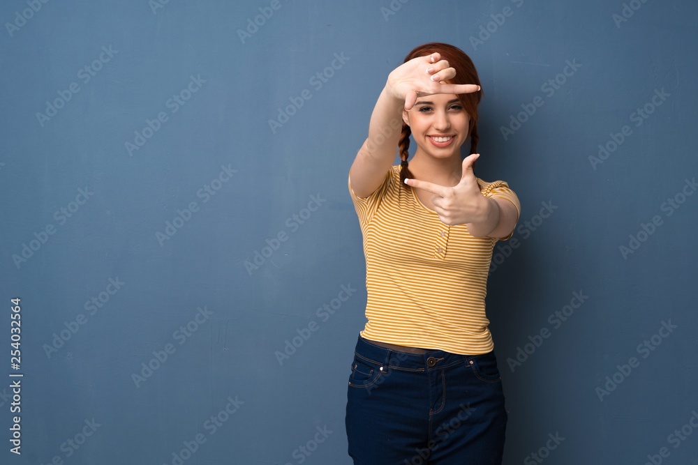 Young redhead woman over blue background focusing face. Framing symbol