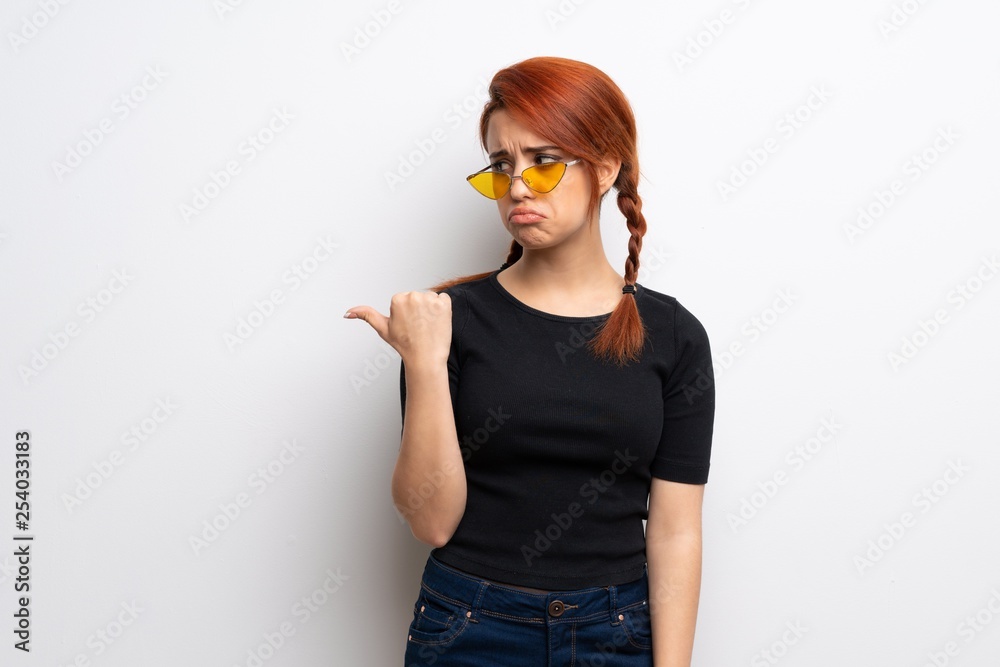Young redhead woman over white wall unhappy and pointing to the side Young redhead woman over white wall unhappy and pointing to the side
